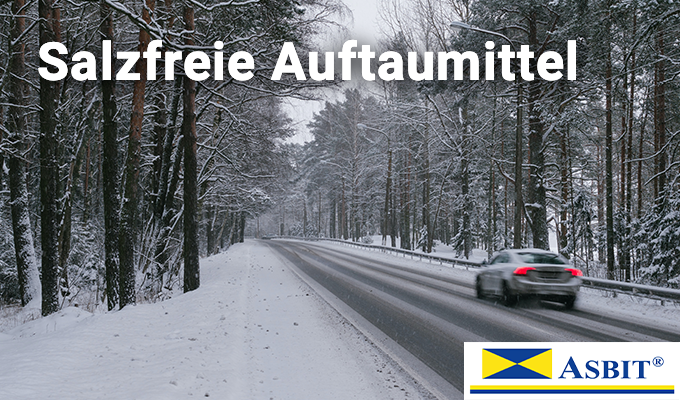 Eine Straße führt durch einen verschneiten Wald, zu beiden Seiten der Straße und auch teilweise auf ihr befindet sich Schnee. Ein graues Fahrzeug fährt gerade auf dem Straßenabschnitt. Salzfreie Auftaumittel verhindern, dass Straßenglätte zu gefährlich wird.
